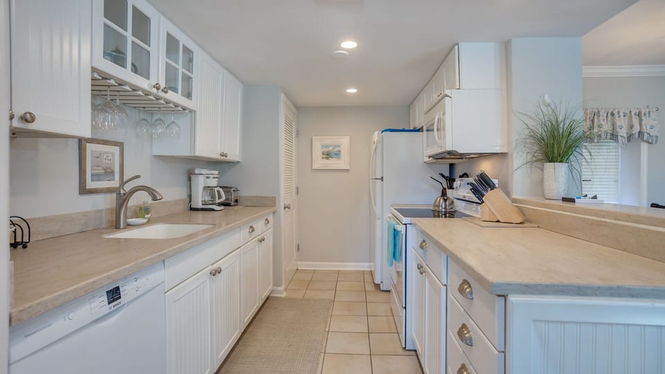 This bright, well-designed galley kitchen offers clean lines and coastal charm with white cabinetry, generous counter space, and recessed lighting throughout.