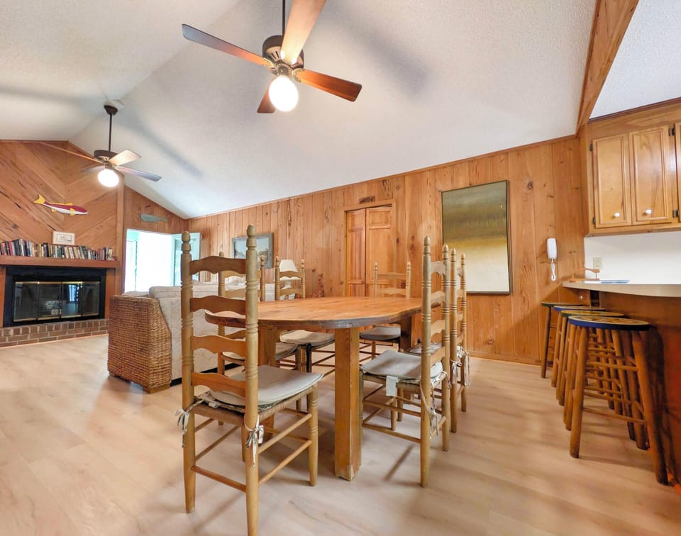 Gather around the handcrafted wooden dining table in this warm, pine-paneled space with vaulted ceilings and ceiling fans. The adjacent breakfast bar offers extra seating, while the cozy fireplace in the living area creates a welcoming retreat.