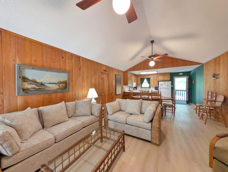 Relax in your spacious, light-filled living area with vaulted ceilings, coastal artwork, and plush sofas. The open layout flows into a fully equipped kitchen and dining space—perfect for gathering after a day at the beach.
