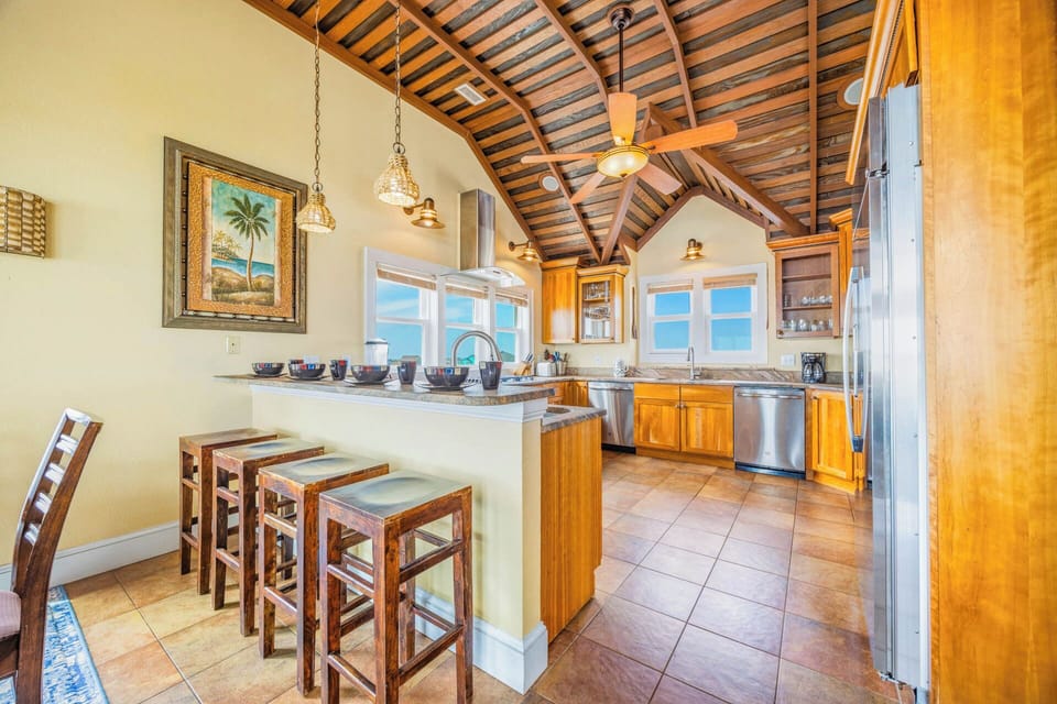Sunlit kitchen with vaulted wood-beam ceilings, an island with bar seating, and ocean views through windows—your perfect space to prepare meals and gather with loved ones.