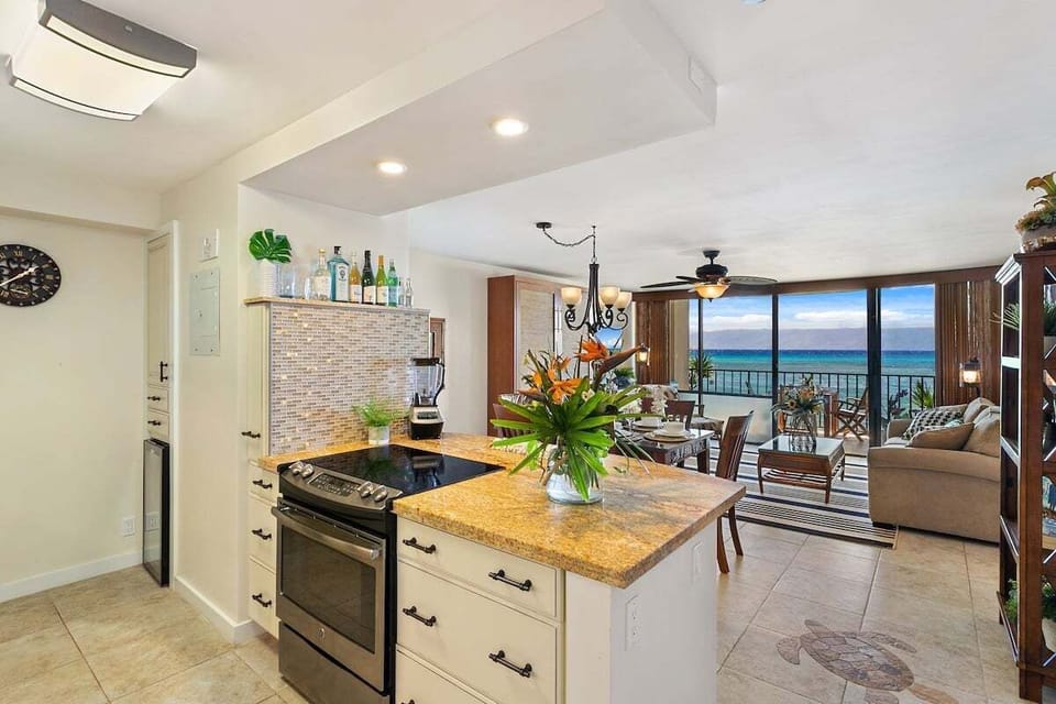 Wide view of kitchen flowing into dining and living areas, tropical flowers and ocean view framed through large windows.
