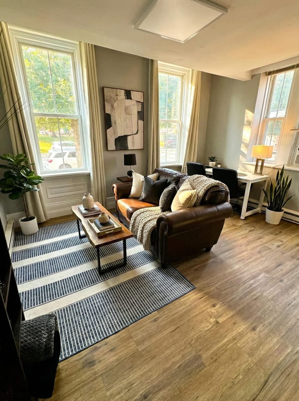 Living area with leather seating, coffee table, TV, tall windows, and workspace nearby.