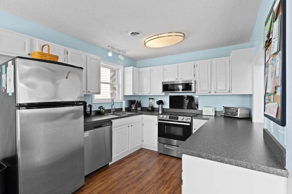 Oak Island, NC kitchen with white cabinets, stainless stove, microwave, fridge, sink, dishwasher, black counters, coffee maker, toaster, and wood floors.