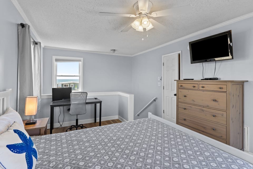Oak Island, NC bedroom with gray bedding, starfish pillow, desk, chair, window ocean view, ceiling fan, wall TV, wood dresser, and coastal style.