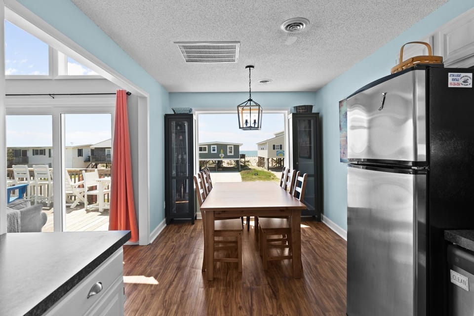 Oak Island, NC dining room with long wood table, chairs, black cabinets, lantern light, blue walls, large picture window, and beach house views.