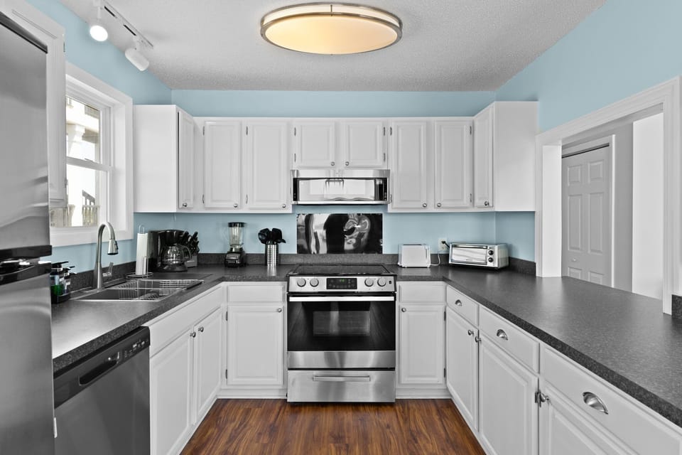 Oak Island, NC kitchen with white cabinets, stainless stove, microwave, fridge, sink, dishwasher, black counters, coffee maker, toaster, and wood floors.