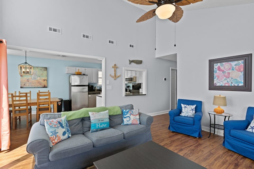 Oak Island, NC living room with gray sofas, blue chairs, TV, coffee table, deck access, ocean views, coral curtains, leaf fan, and high windows.