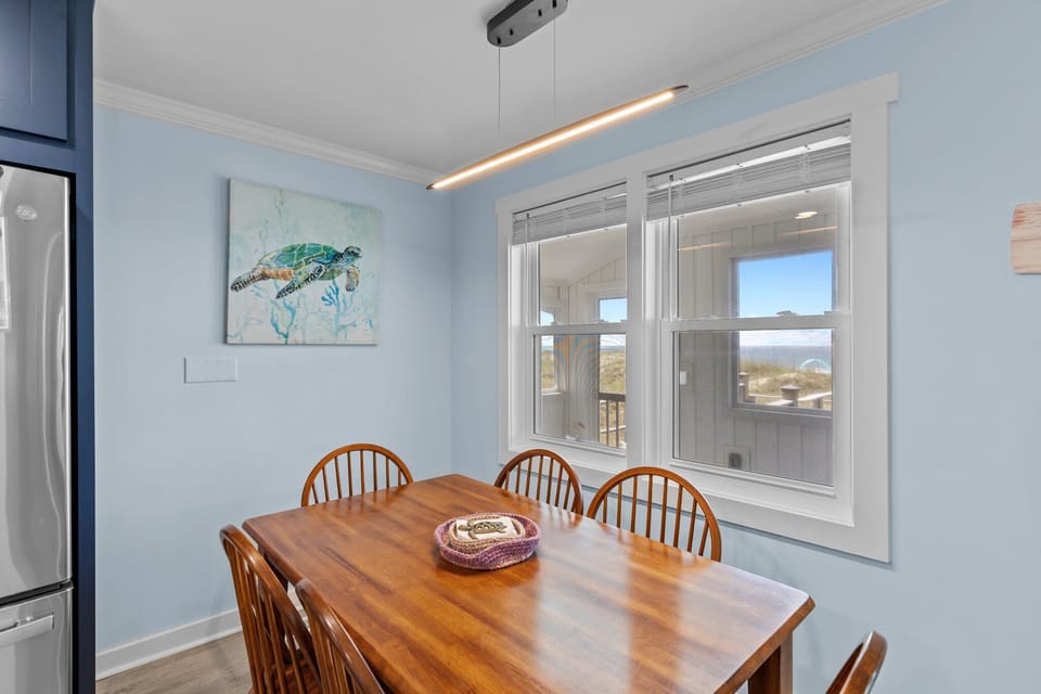 Coastal dining nook with wood table, six chairs, turtle wall art, modern light fixture, blue walls, white-trim windows, and ocean views.