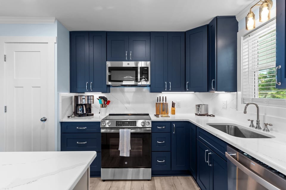 Bright kitchen with navy cabinets, white island, stainless appliances, pendant lights, marble counters, window over sink, pantry door, and coastal decor.