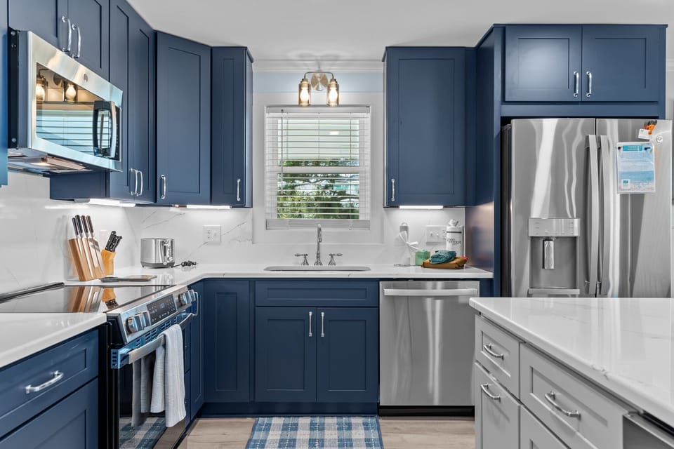 Bright kitchen with navy cabinets, white island, stainless appliances, pendant lights, marble counters, window over sink, pantry door, and coastal decor.