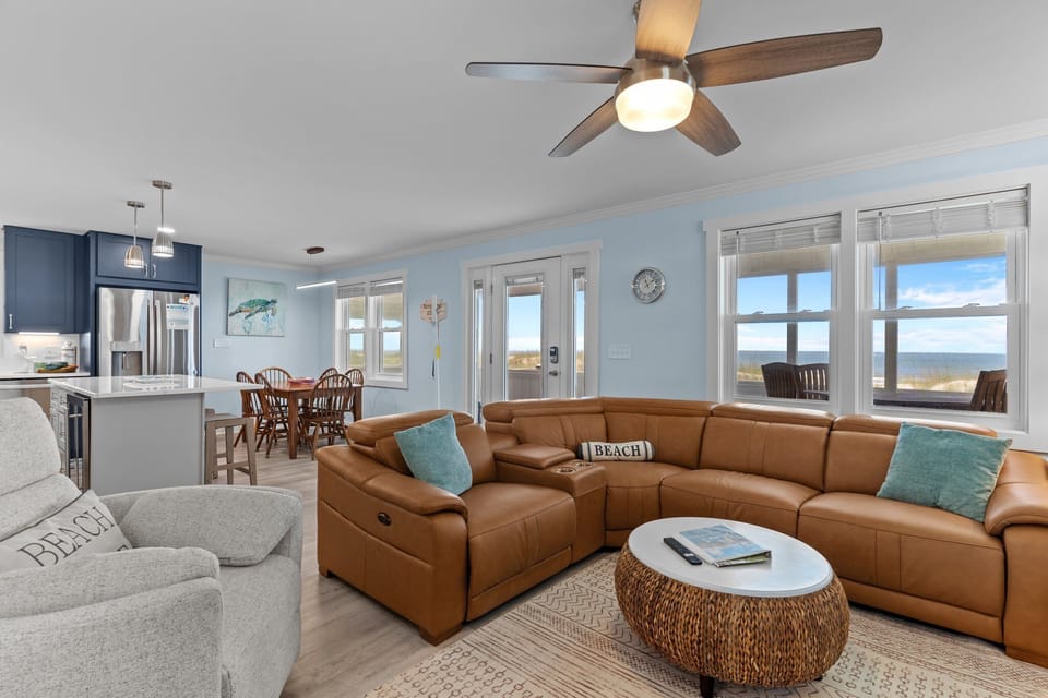 Bright living room with leather sectional, recliner, wall TV, ceiling fan, bookshelves, lamps, beach art, round coffee table, and light blue walls.