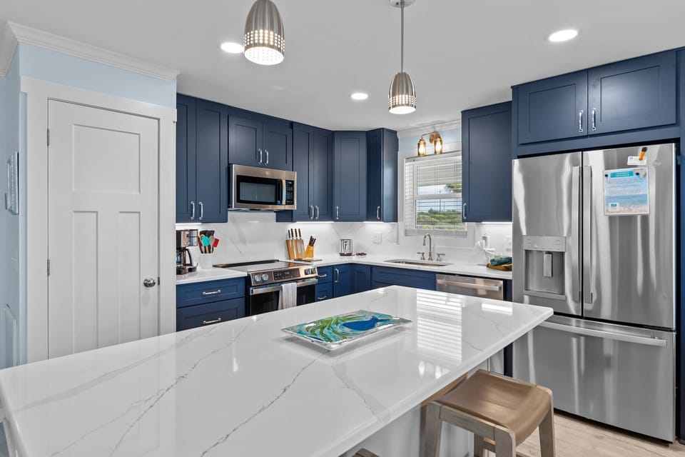 Bright kitchen with navy cabinets, white island, stainless appliances, pendant lights, marble counters, window over sink, pantry door, and coastal decor.