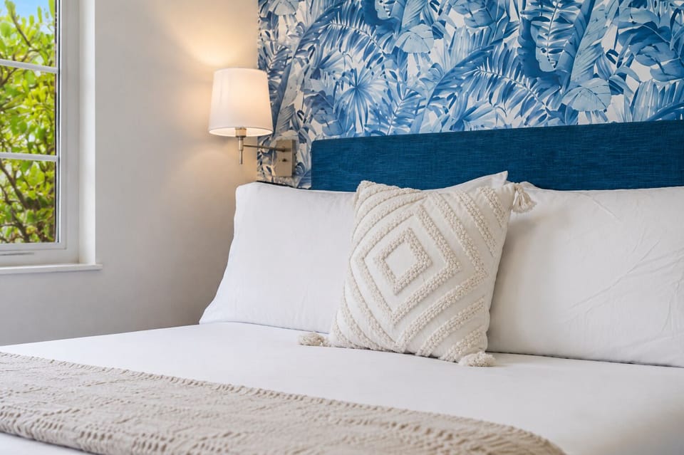Bold blue palm-print wallpaper backs the headboard here, with a woven diamond pillow and soft white linens keeping the look breezy and beach-ready. A wall sconce close by makes late-night reading easy without leaving the bed.