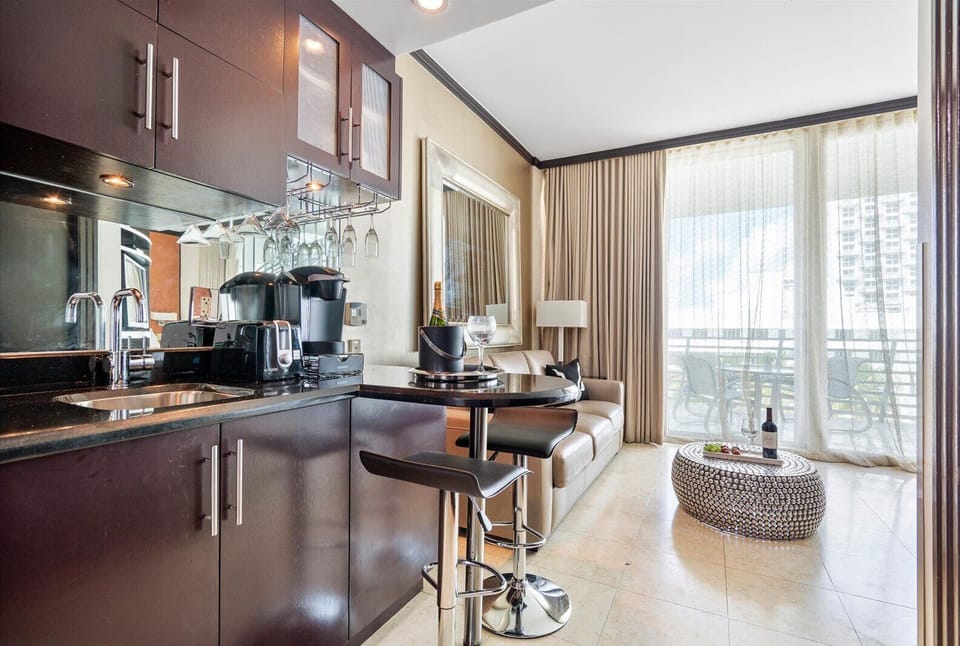 Compact modern kitchen with dark cabinetry and a small seating area featuring a barstool and a sofa. Floor-to-ceiling windows illuminate the space, leading to a balcony with outdoor seating.