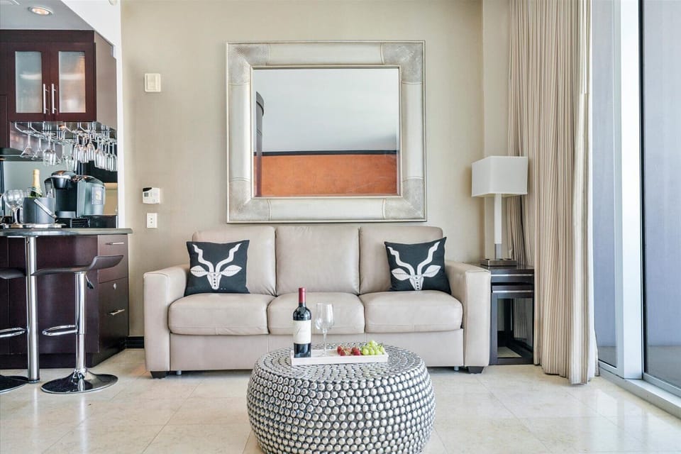 Modern living room with a beige couch, fox-themed pillows, a decorative mirror on the wall, a round patterned coffee table holding a wine bottle and grapes, and floor-to-ceiling windows with curtains.
