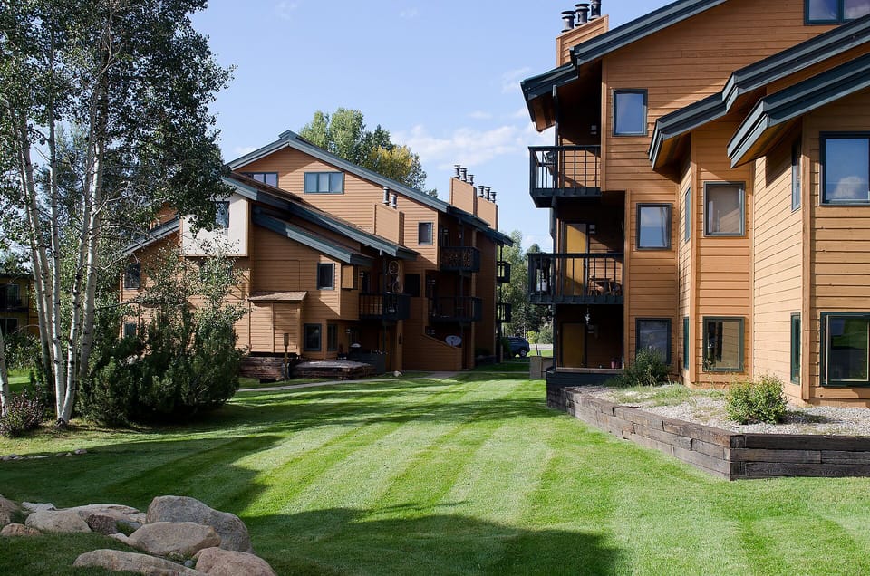 Mountain resort buildings featuring warm wood architecture set among mature trees and manicured lawns with scenic balconies.