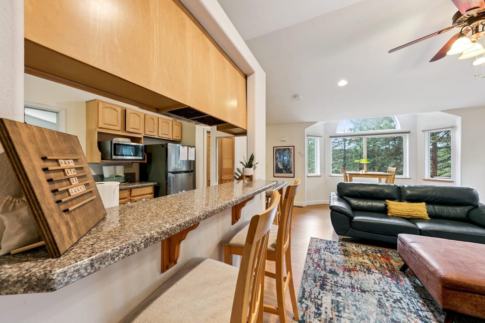 Granite breakfast bar with wooden stools overlooks the kitchen and living area with black leather couch and colorful rug.