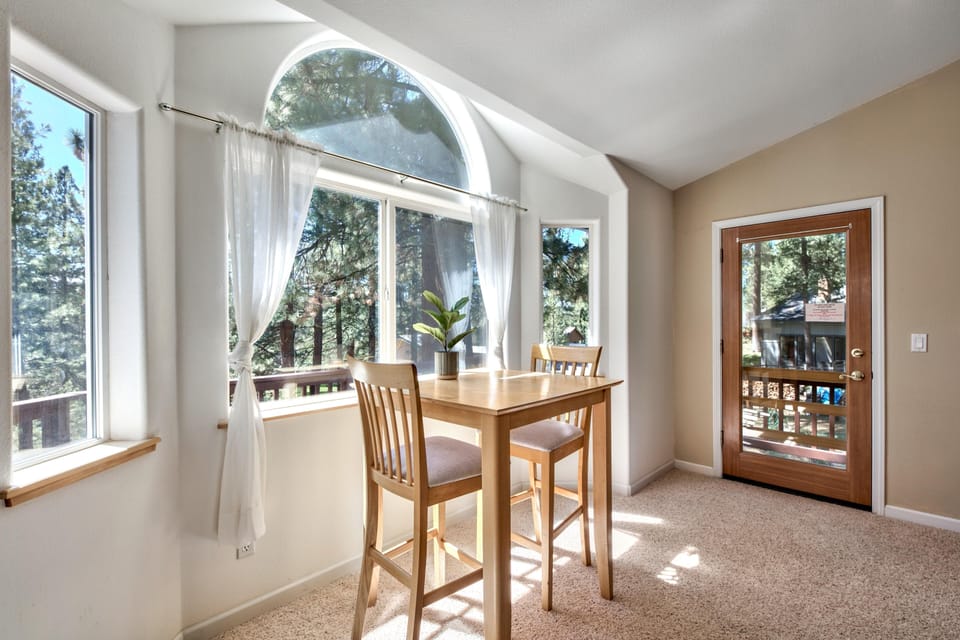 Breakfast nook with light wood table and chairs beside arched window and glass door overlooking pine trees.