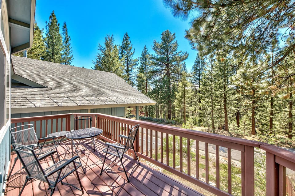 Wooden balcony with metal mesh chairs and round table overlooks tall pine trees under clear blue sky.