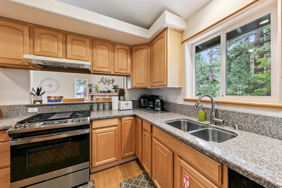 Kitchen corner with stainless steel gas range, granite counters, light wood cabinets, and a window overlooking pine trees.