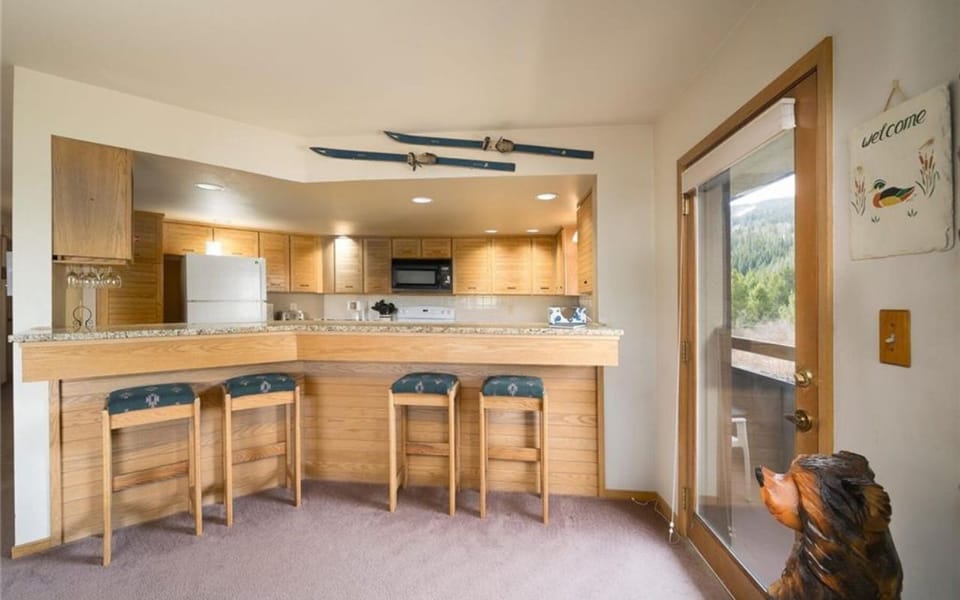 A kitchen with wooden cabinets, a breakfast bar with three stools, and a white refrigerator. Above the bar hangs a pair of skis. The glass door on the right leads to an outdoor view of trees.
