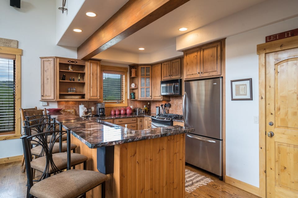 A kitchen with wooden cabinets, stainless steel appliances, a marble countertop, and four bar stools at an island. Bright natural light filters through the windows.