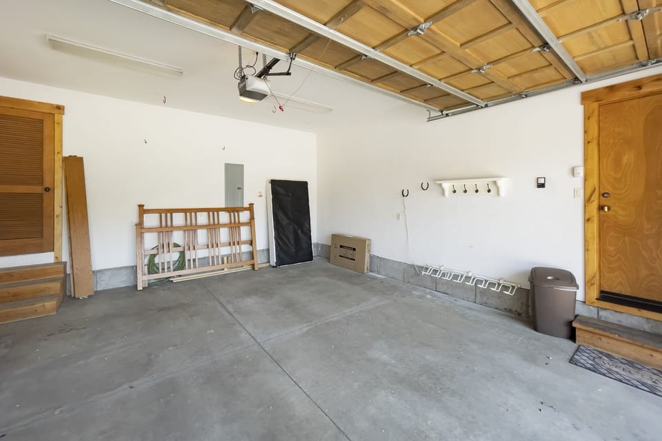 Empty garage with wooden steps, a closed wooden door, a recycling bin, a small shelving unit, and a few miscellaneous items like a baby gate and a bicycle.