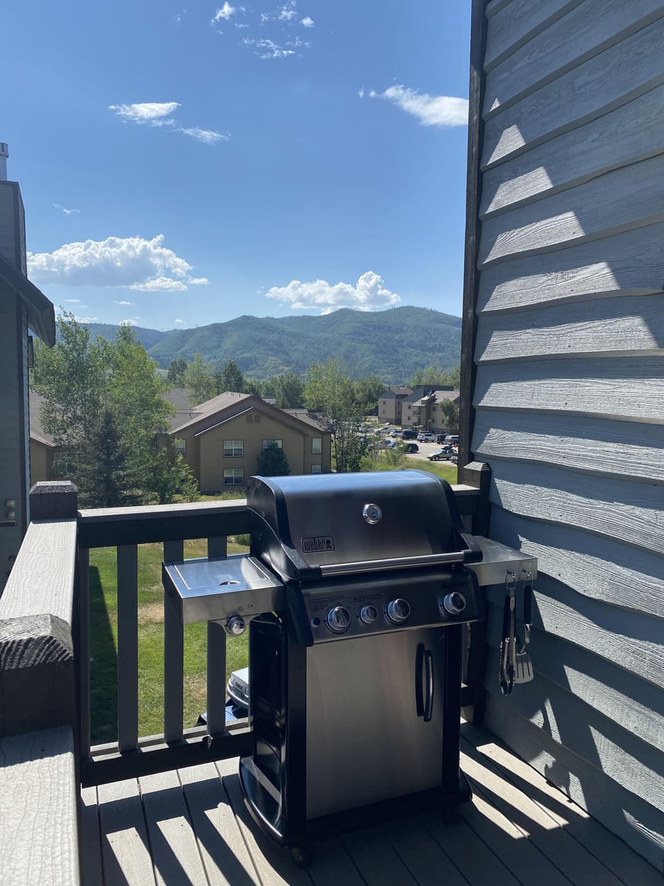 A propane grill sits on a wooden deck overlooking a residential area with mountains in the background under a partly cloudy sky.