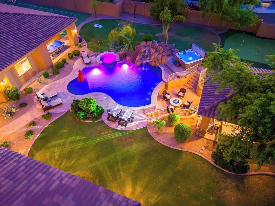 Aerial view of a resort-style property featuring a custom pool with illuminated grotto, elevated hot tub, multi-level terraces with lounge seating, and lush landscaping throughout the expansive backyard oasis.