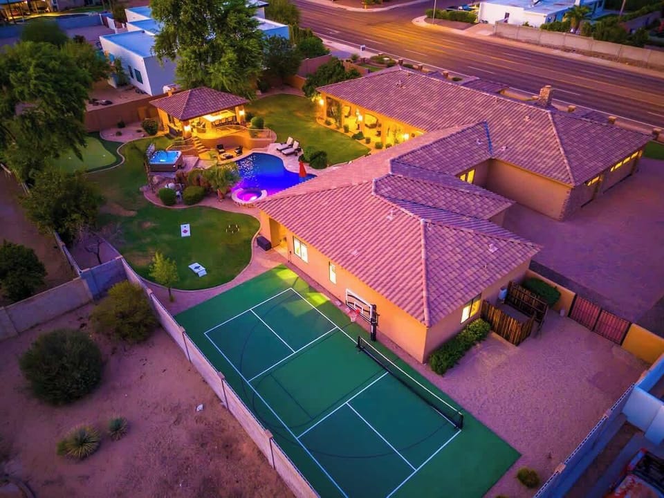 Aerial view of an expansive estate at dusk featuring a tennis court, pool with hot tub, manicured lawns, and multiple outdoor entertainment areas across a sprawling property.