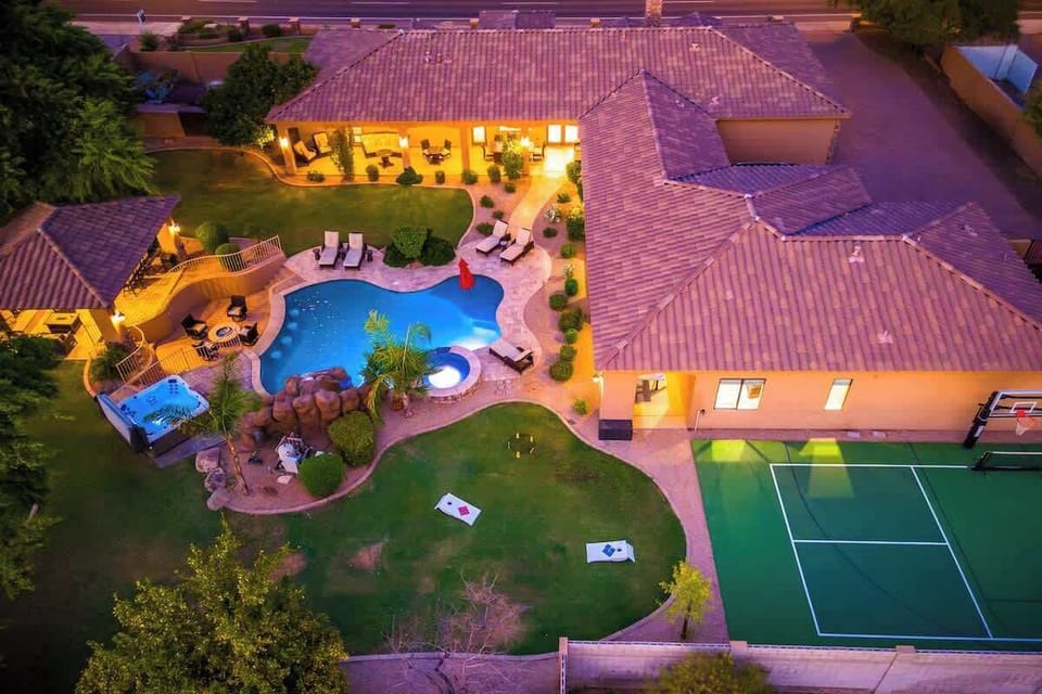 Aerial view of the property at dusk, showcasing a resort-style pool, tennis court, spacious lawns, and covered patio areas surrounded by desert landscaping.