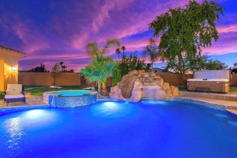 A stunning backyard oasis awaits at dusk, where a glowing pool with a rock waterfall feature and adjacent hot tub invite relaxation beneath vibrant desert skies painted in shades of purple and blue.