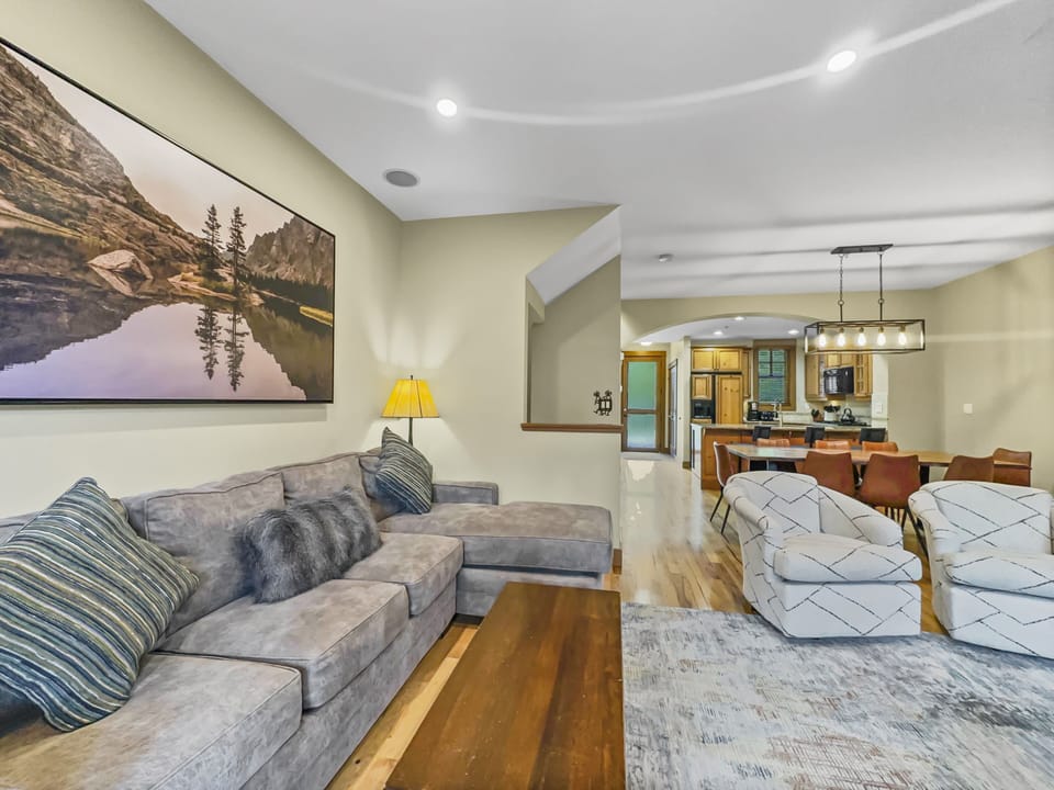 A living room with a gray sectional sofa, patterned chairs, wooden coffee table, wall art of a lake scene, and a view into a kitchen and dining area with a light fixture and brown dining chairs.