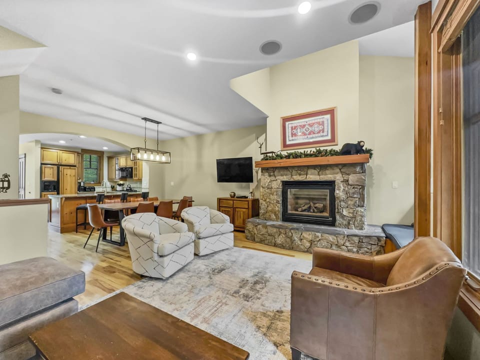 Living room with stone fireplace, TV mounted above, two armchairs, a leather chair, and a coffee table. Open concept kitchen and dining area with modern lighting visible in the background.