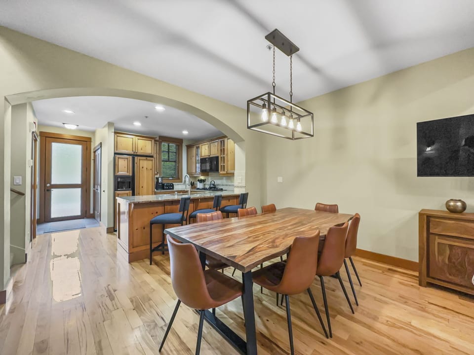 A dining room with a wooden table and six brown chairs. A kitchen with wooden cabinets is visible through an arched doorway. A pendant light hangs above the table, and a sideboard is against the wall.