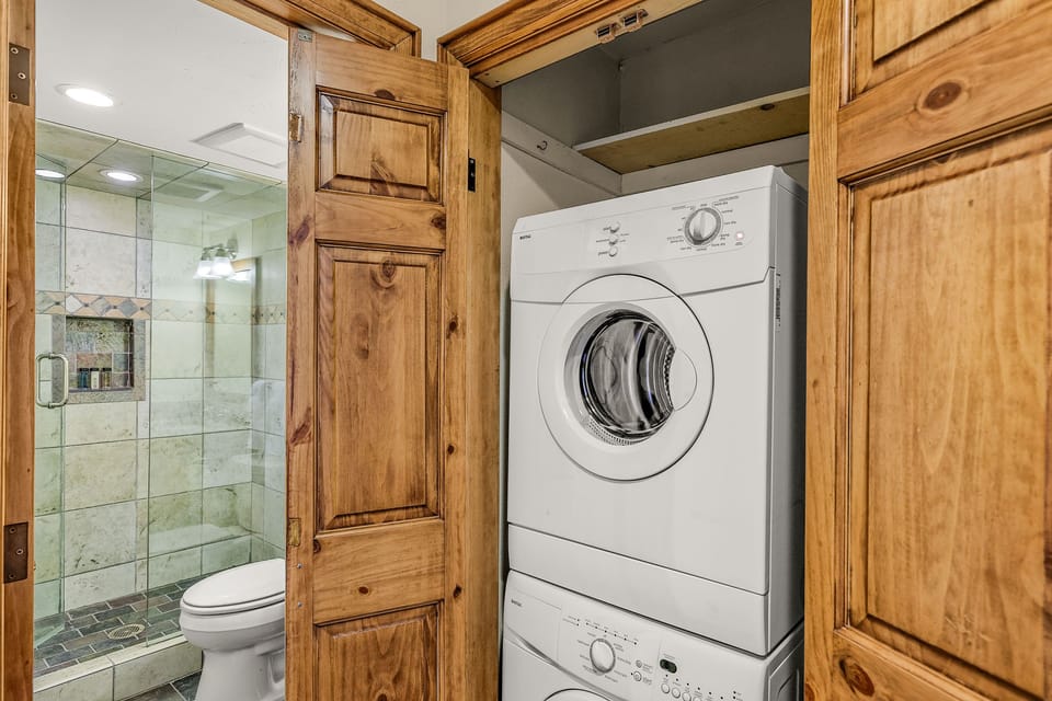 A laundry area with a stacked washer and dryer is seen in a wooden closet next to a bathroom with a glass-enclosed shower and a toilet.