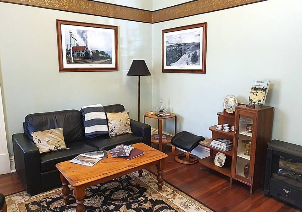 This inviting living room features a black leather sofa with patterned cushions, a wooden coffee table, and a cozy armchair with a matching footrest. Decor includes framed vintage photographs, a small bookcase with teacups and memorabilia, and a floor lam