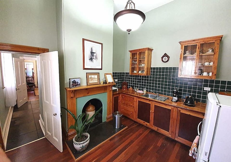A charming vintage-style kitchen features rich wooden cabinetry, green tiled backsplash, and a decorative fireplace with framed photos above. The space includes a sink, modern appliances, and a mix of classic and cozy decor under a high ceiling with a cen