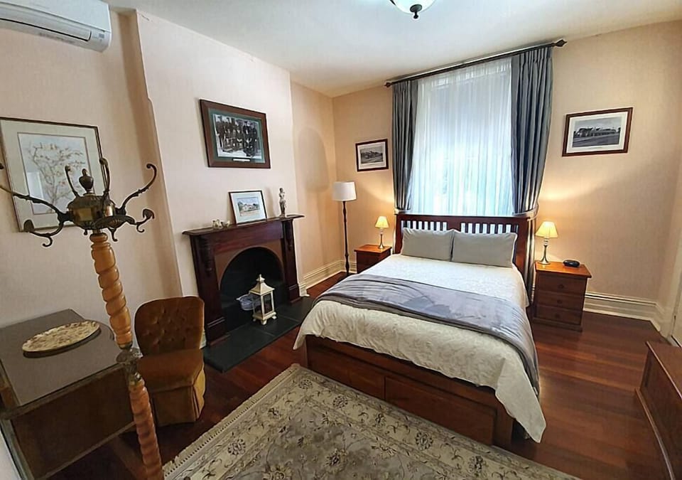 This cozy, vintage-style bedroom features a wooden-framed double bed with elegant bedding, matching bedside tables with lamps, and a classic fireplace. The room is softly lit with natural light through sheer curtains, complemented by framed artwork and wa