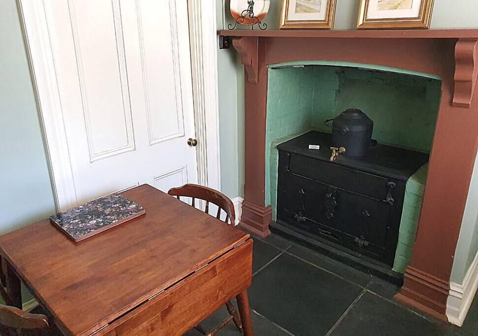 This cozy corner features a small wooden dining table with two matching chairs, placed beside a vintage cast iron stove set into a painted brick fireplace. The hearth is accented with a muted red-brown mantel and soft green interior walls, adding a rustic
