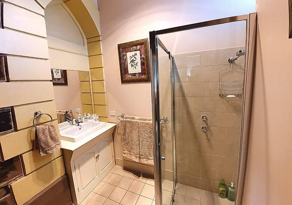 This charming bathroom features a glass-enclosed walk-in shower with beige tiles, a classic white vanity with twin faucets, and a large mirror framed by decorative archways. Soft brown towels and floral artwork on the walls add a warm, welcoming touch to 