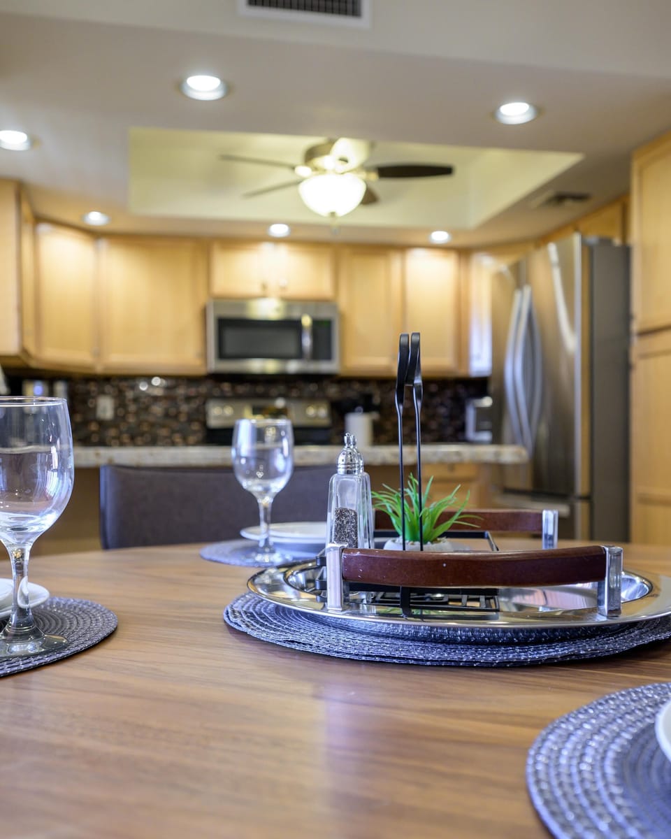 Wood dining table set with glassware and placemats, overlooking light wood kitchen cabinets and stainless appliances.