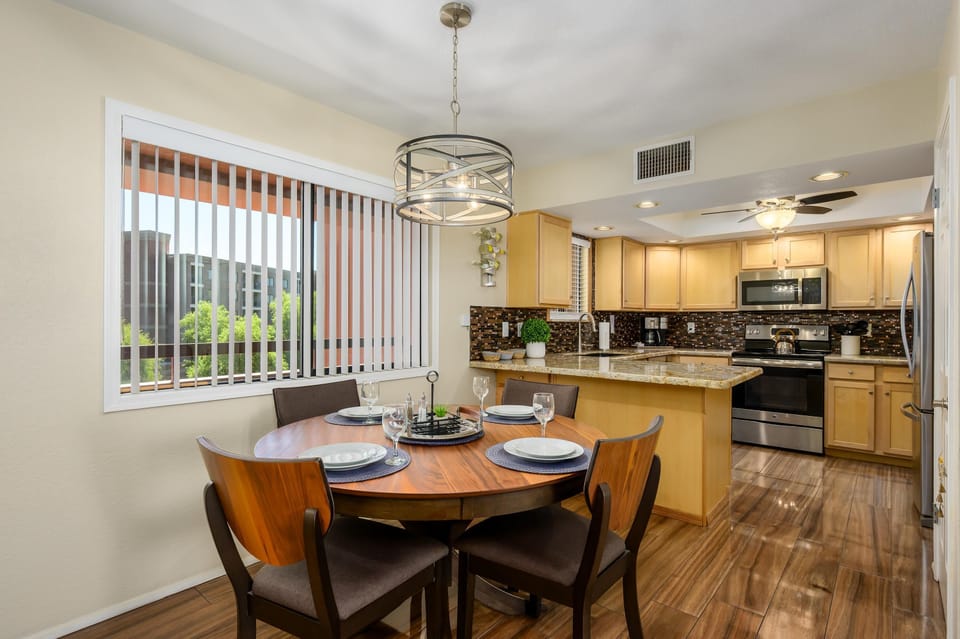 Round wooden dining table set with four chairs sits beside a large window with vertical blinds and a modern metal chandelier.