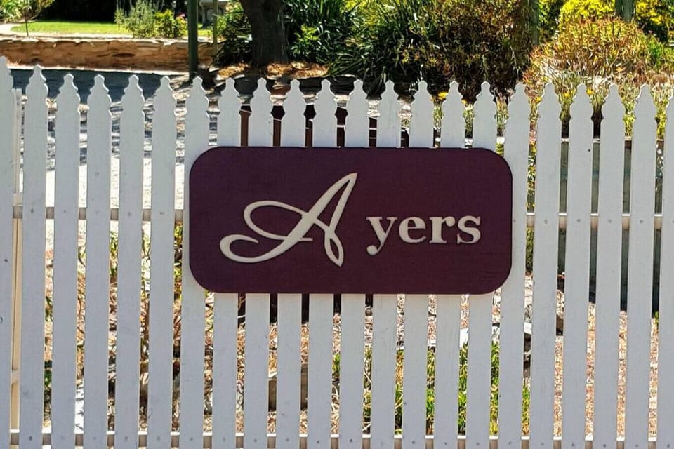 A white picket fence features a maroon sign with the word “Ayers” in elegant white lettering. The decorative “A” stands out in a stylish script, giving a charming and personalized touch to the entrance.
