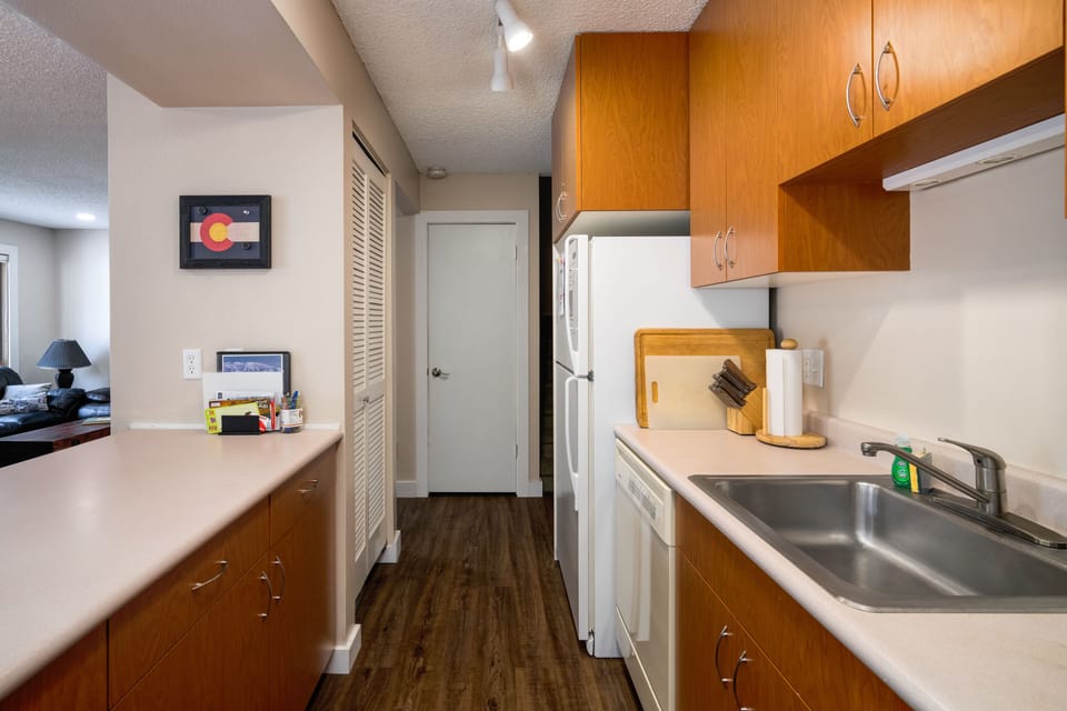 A compact kitchen with wooden cabinets, white appliances, and beige countertops. Adjacent to the kitchen is a living area with a black lamp and a couch. An artwork is mounted on a wall near the kitchen.