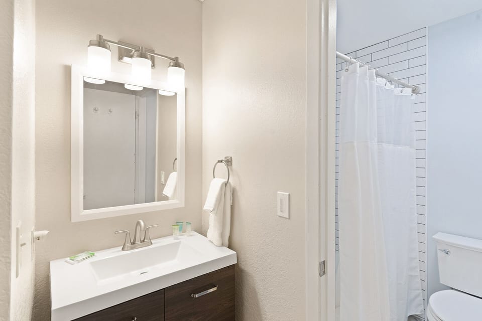 A modern bathroom features a vanity with a mirror, sink, and light fixture. A white towel is hanging on a wall-mounted towel ring next to the mirror. A shower with a white curtain is visible in the background.