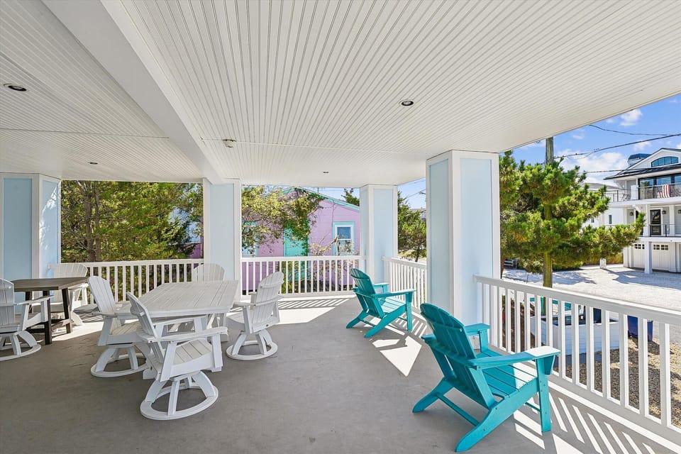 A spacious covered porch features turquoise Adirondack chairs and a white dining set, offering a bright, coastal retreat with neighborhood views.
