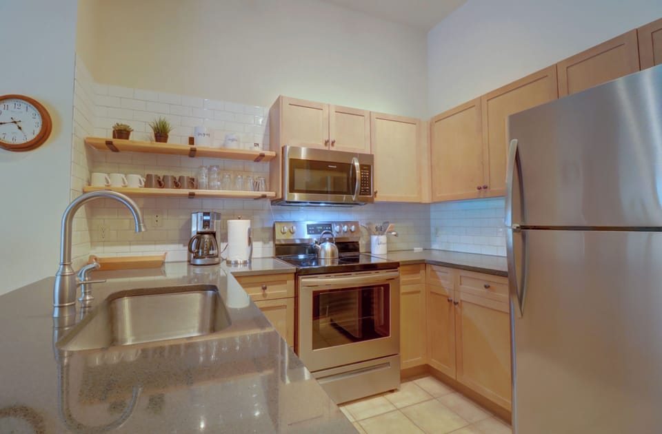 A modern kitchen with stainless steel appliances, wooden cabinets, a sink, and an electric stove. Shelves above the counter hold dishes and plants. A round clock is on the wall.
