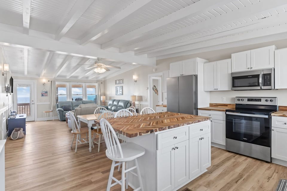 Bright kitchen with white cabinetry, stainless appliances, butcher block counters, and island seating for four, plus coffee maker and window views in this Oak Island, NC beach home.