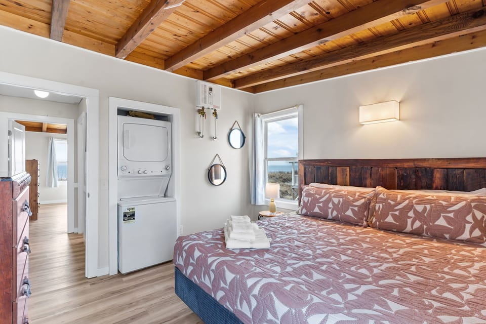 Comfortable bedroom with wood beam ceiling, queen bed, dresser with mounted TV, window for natural light, and coastal décor in this Oak Island, NC beach home.